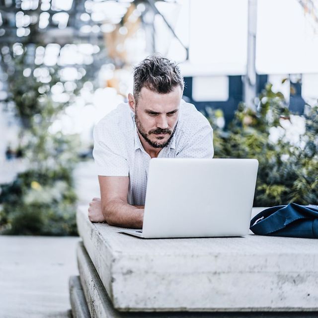 Person arbeitet im Freien liegend mit einem Laptop auf einer Betonkante, im Hintergrund grüne Pflanzen.