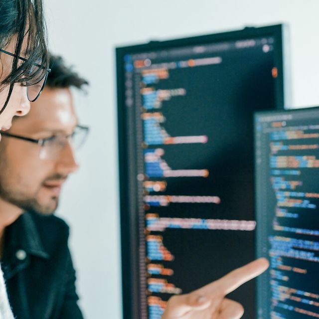 Zwei Personen analysieren Quellcode auf großen Monitoren, eine zeigt mit dem Finger auf den Bildschirm.