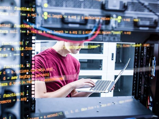 Person im Rechenzentrum arbeitet mit einem Laptop zwischen Serverschränken, überlagert von Programmcode auf dem Bildschirm.