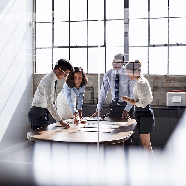 Arbeitsgruppe im modernen Büro steht um einen Tisch und bespricht Unterlagen, durch Glaswand fotografiert
