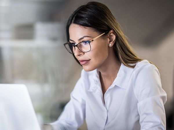 Frau mit Brille und weißer Bluse arbeitet konzentriert am Laptop im Büro, Notizbuch und Stift liegen daneben.