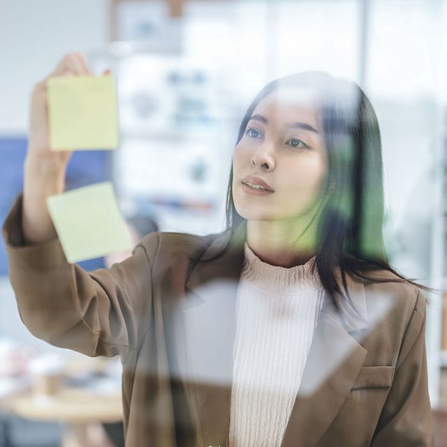 Frau im Business-Blazer klebt gelbe Haftnotizen an eine Glaswand im Büro.