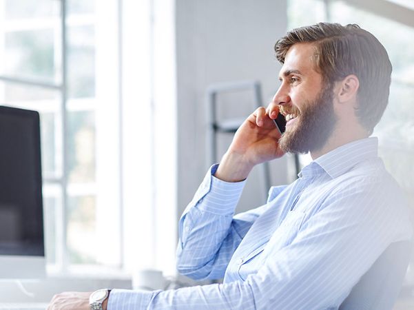 Lächelnder Mann im blauen Hemd telefoniert mit dem Smartphone am Schreibtisch in einem hellen Büro.