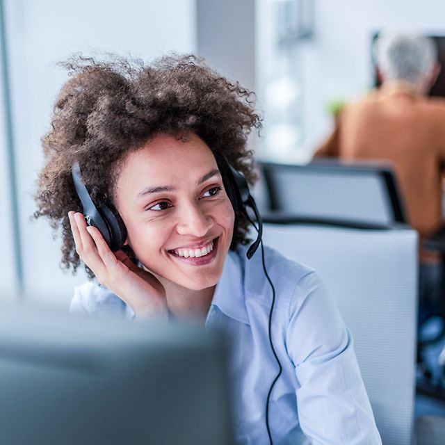 Lächelnde Frau mit Headset und lockigem Haar im Büro, blickt zur Seite; im Hintergrund arbeitet ein Kollege am PC.