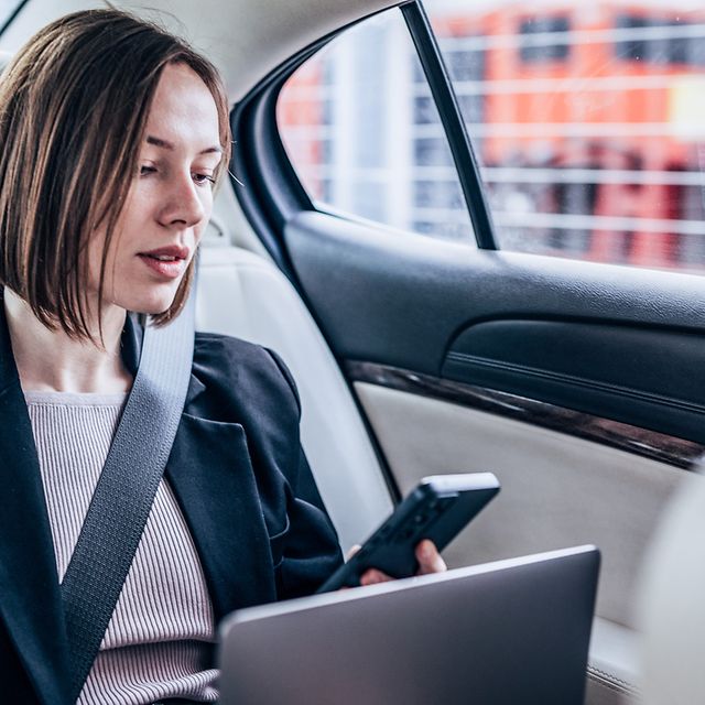Frau sitzt mit ihrem Laptop und Smartphone im Auto und schaut auf den Bildschirm.
