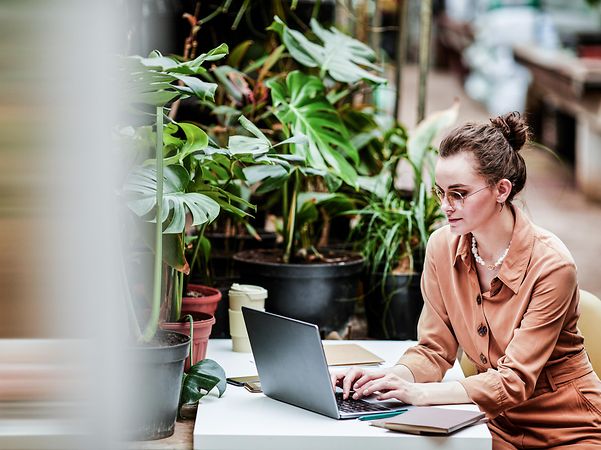 Eine Frau sitzt vor Pflanzen und schaut auf ihren Laptop.