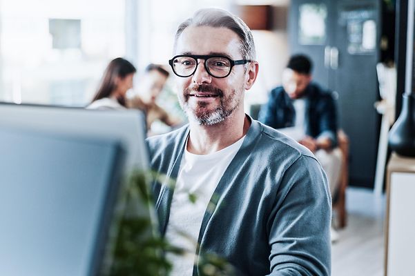 Mann im Büro am Computer, im Hintergrund ein Teammeeting